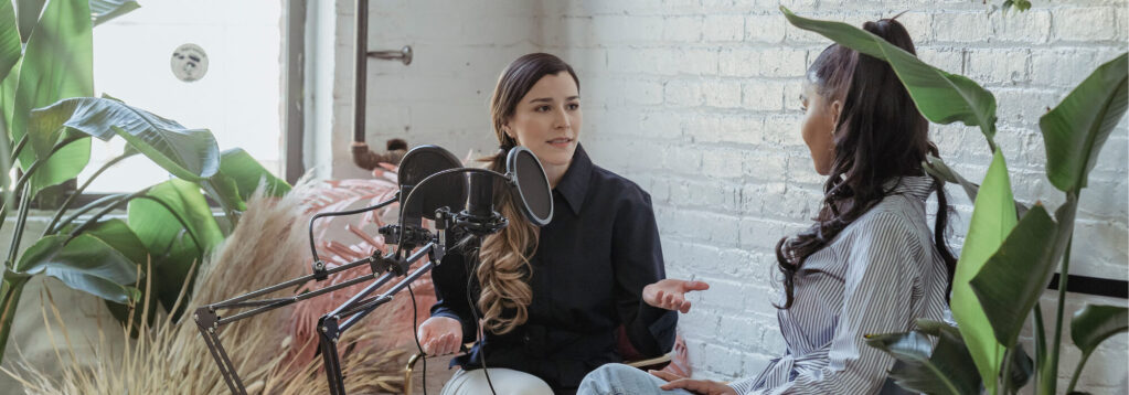 Two women with long hair speaking into microphones livestreaming during a podcast, sitting indoors by a white brick wall, gesturing with one hand during a recording session. 3D rendering, design studio, 3D product, animation, character design, 2D animation, video production services, book trailer services, digital marketing services, motion design, motion graphics. RDV Media – Multimedia production company. Serving brands globally. New York, Los Angeles, California, Atlanta, USA, Dublin, Ireland, EU, London, England, UK, Doha, Qatar, UAE, Abu Dhabi, Ajman, Dubai, Fujairah, Ras Al Khaimah, Sharjah, and Umm Al Quwain.