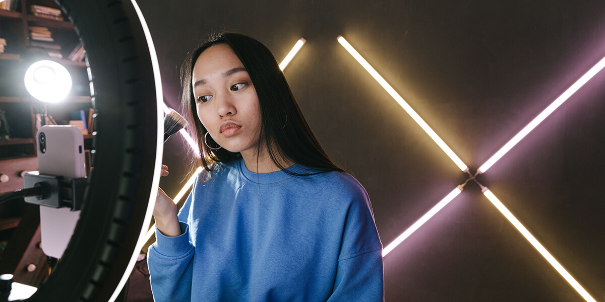 Woman in a blue sweatshirt holding a makeup brush, looking at a mirror, with neon lights in the background.