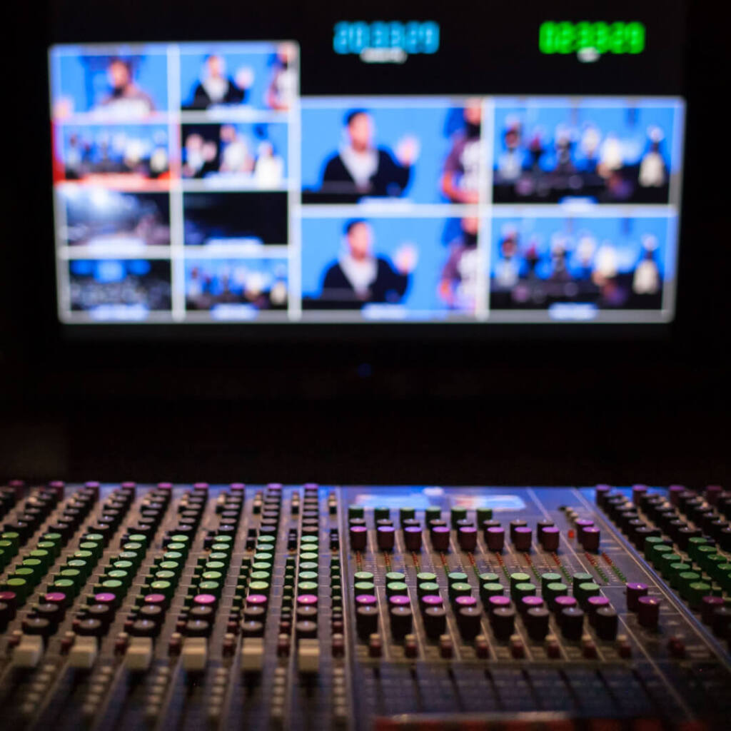 Audio mixing console with illuminated buttons in the foreground, broadcast control screens showing multiple video feeds in the background. 3D rendering, design studio, 3D product, animation, character design, 2D animation, video production services, book trailer services, digital marketing services, motion design, motion graphics. RDV Media – Multimedia production company. Serving brands globally. New York, Los Angeles, California, Atlanta, USA, Dublin, Ireland, EU, London, England, UK, Doha, Qatar, UAE, Abu Dhabi, Ajman, Dubai, Fujairah, Ras Al Khaimah, Sharjah, and Umm Al Quwain.