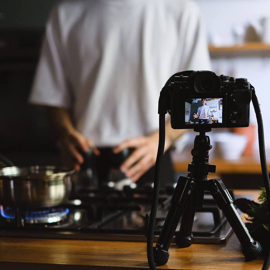 Person filming a cooking tutorial in a kitchen, standing behind a camera on a tripod with a pot on a stove. 3D rendering, design studio, 3D product, animation, character design, 2D animation, video production services, book trailer services, digital marketing services, motion design, motion graphics. RDV Media – Multimedia production company. Serving brands globally. New York, Los Angeles, California, Atlanta, USA, Dublin, Ireland, EU, London, England, UK, Doha, Qatar, UAE, Abu Dhabi, Ajman, Dubai, Fujairah, Ras Al Khaimah, Sharjah, and Umm Al Quwain.