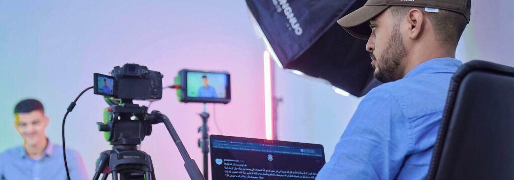 A YouTube livestream recording studio setup with camera equipment, softbox lighting, a laptop displaying a script and live comments, and a blurred screen in the background showing a person active on stream. RDV Media – Multimedia production and marketing company. Serving brands globally.