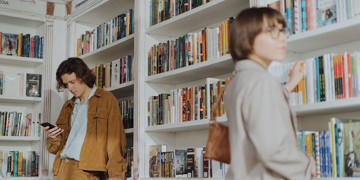 Two people browsing bookshelves in a modern library, filled with colorful books; one person wearing a brown jacket is watching a book trailer on their cellphone and the other carrying a tan handbag. RDV Media – Multimedia production company. Serving brands globally.