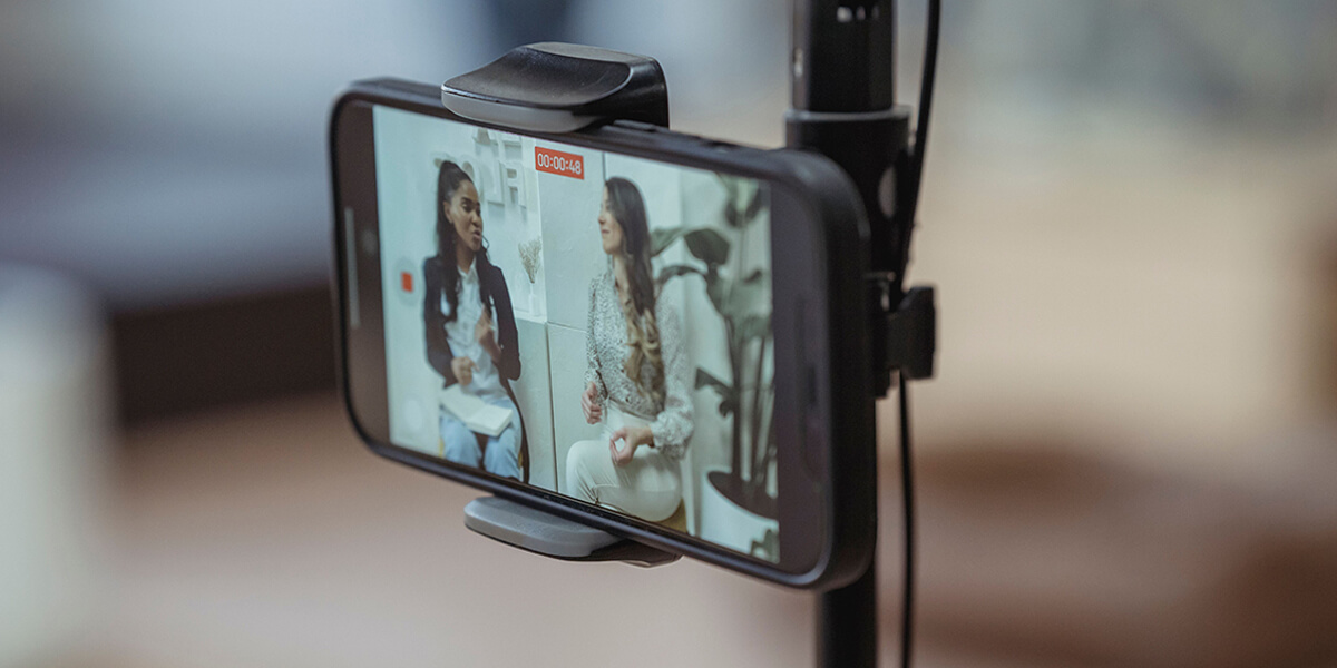 Two women having a conversation recorded on a smartphone, mounted on a tripod, for a YouTube livestream, with a studio background and recording interface visible on the screen. RDV Media – Multimedia production company. Serving brands globally.