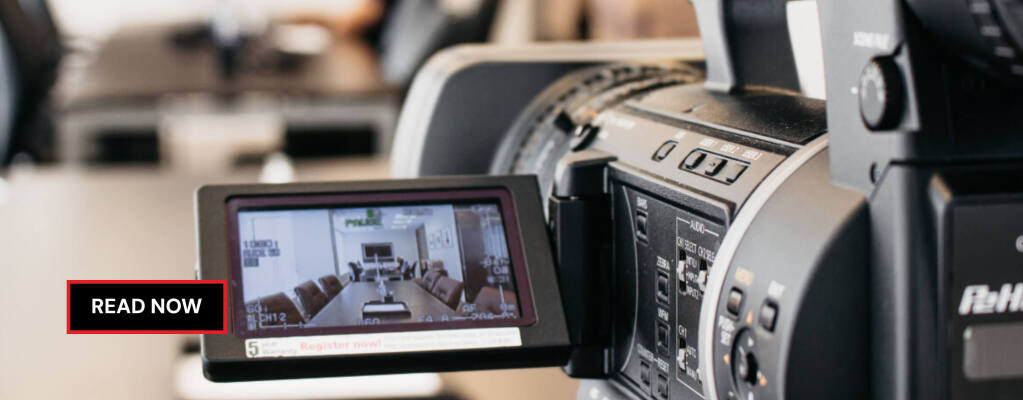 Video camera recording a conference room setup, with a blurred person in the background and the scene displayed on the camera's LCD screen. RDV Media – multimedia production and marketing company. Serving brands globally.