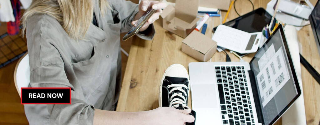 Woman using an iPhone to capture video of a sneaker near a laptop and cardboard boxes on a wooden desk, creating product videos for online selling. RDV Media – multimedia production and marketing company. Serving brands globally.