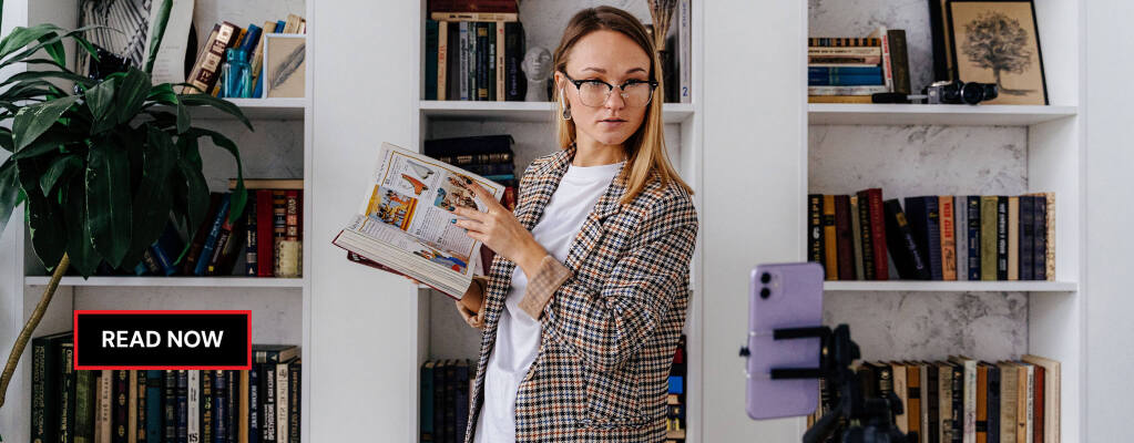 Woman wearing glasses holding an open book, standing by a bookshelf, looking towards a smartphone on a tripod, recording a video of herself for social media. RDV Media – multimedia production and marketing company. Elevated content for a global audience.