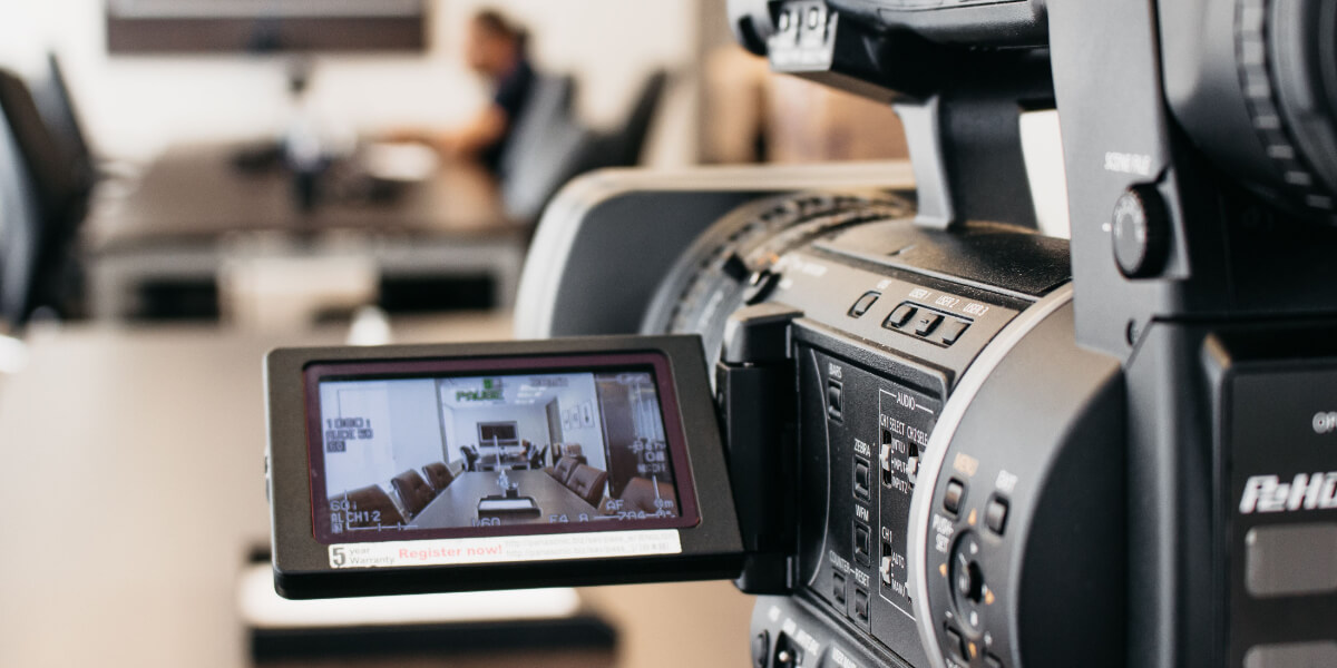 Video camera recording a conference room setup, with a blurred person in the background and the scene displayed on the camera's LCD screen. RDV Media – multimedia production and marketing company. Serving brands globally.