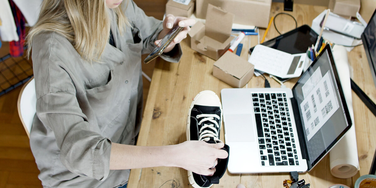 Woman using an iPhone to capture video of a sneaker near a laptop and cardboard boxes on a wooden desk, creating product videos for online selling. RDV Media – multimedia production and marketing company. Serving brands globally.